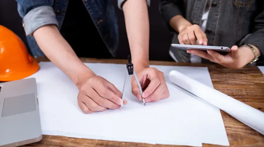 Hands using a compass on blueprint paper with a laptop and construction helmet nearby, illustrating construction planning and permitting support for landscaping projects.