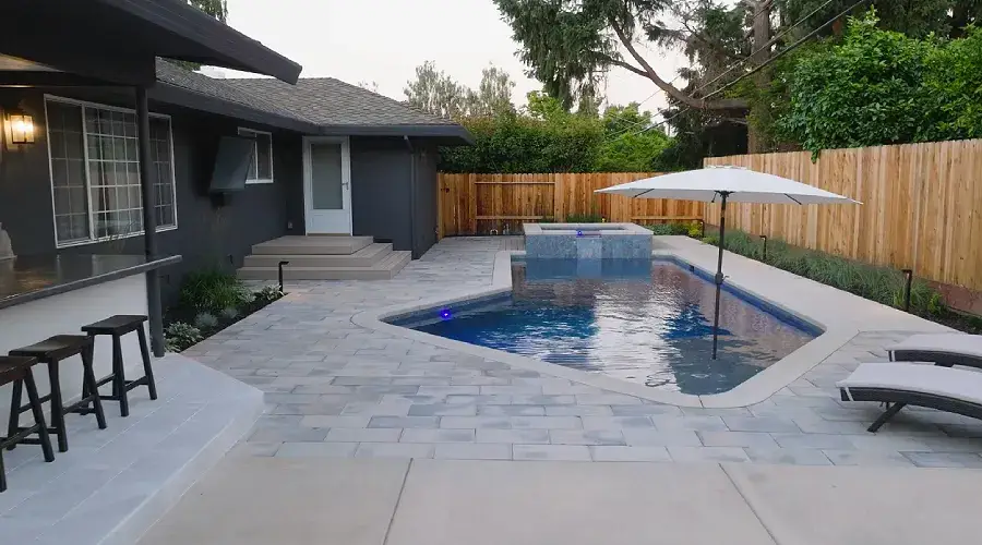Paver patio with poolside lounging area, featuring a modern design, surrounded by greenery and wooden fencing, enhancing outdoor living space.