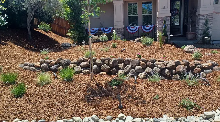 Rock retaining wall with natural stone, surrounded by mulch and various plants in a landscaped yard, enhancing outdoor aesthetics and functionality.
