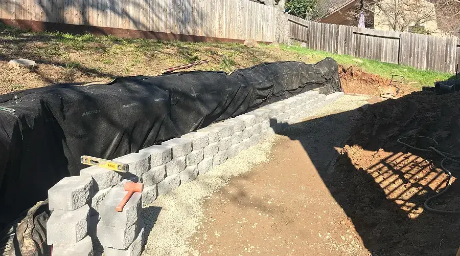 Split-face stone retaining wall under construction, showcasing premium craftsmanship and quality materials, with tools and landscape fabric visible, emphasizing Rogers Landscape's commitment to durable and aesthetic landscaping solutions.