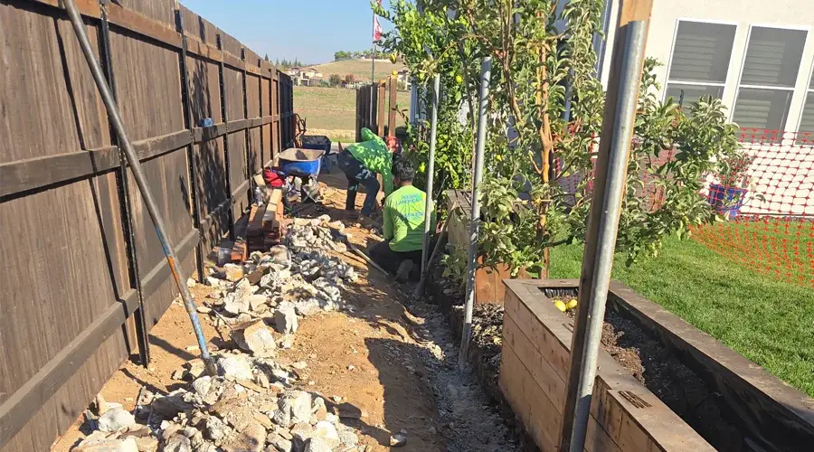 Team of workers removing concrete debris in a residential yard, ensuring careful demolition and site preparation for landscaping, with safety measures in place and greenery visible.