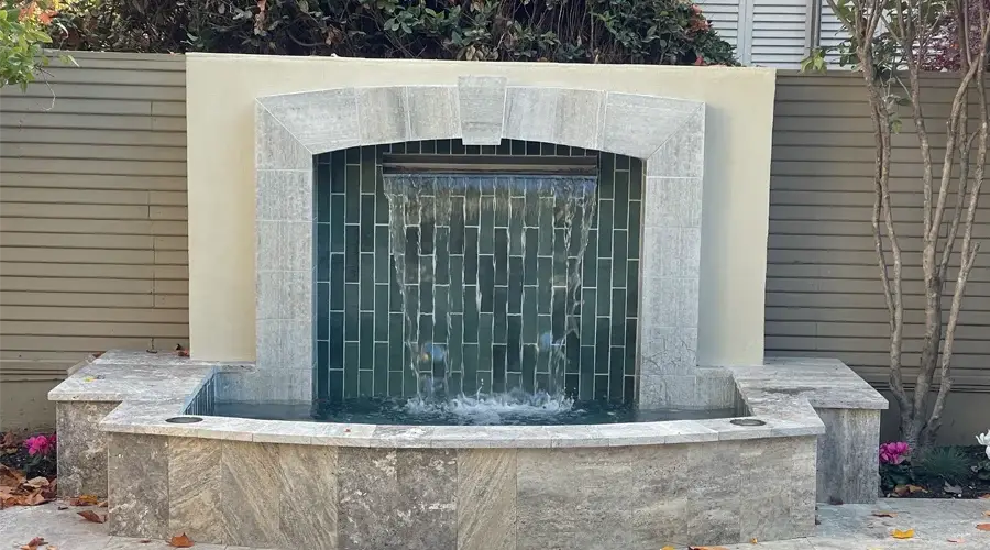 Custom water wall with flowing water feature, surrounded by stone seating and green tiles, enhancing outdoor landscape aesthetics.