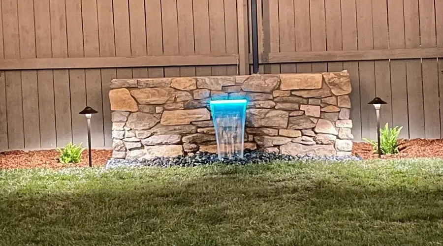 Custom water wall with illuminated blue flow, surrounded by stone and landscaping, enhancing outdoor serenity and design.