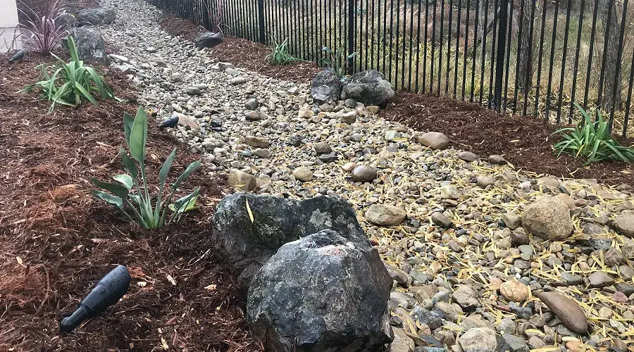 Decorative rock creek bed landscaping with various sizes of rocks, surrounded by mulch and green plants, enhancing outdoor aesthetics and functionality.