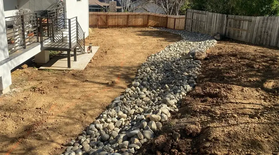 Drainage system installation in Loomis, CA, featuring a rock-lined channel for water management, with a partially cleared yard and a wooden fence in the background.