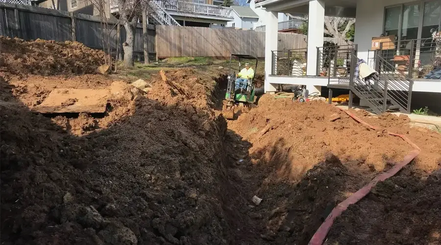 Excavation site for drainage installation featuring a worker operating machinery in a trench, surrounded by freshly dug soil and residential structures, illustrating Rogers Landscape's drainage solutions in action.