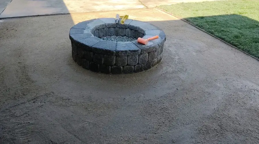 Custom stone fire pit installation in a landscaped outdoor setting, featuring gravel interior and tools for construction, emphasizing Rogers Landscape's outdoor living services.