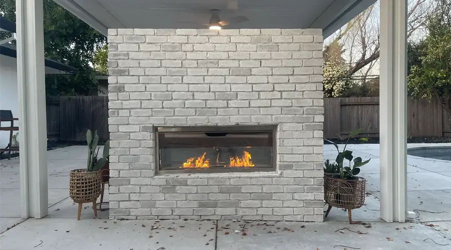 Modern outdoor gas fireplace with a white brick surround, featuring a glass front and flames visible, surrounded by potted plants in a backyard setting.