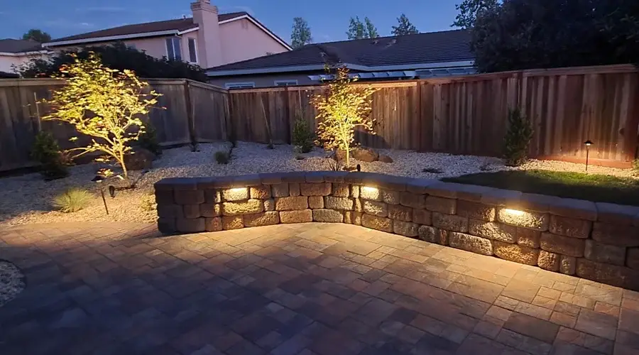 Outdoor landscape lighting illuminating a stone wall and surrounding trees in a residential backyard, enhancing safety and aesthetic appeal for evening outdoor enjoyment.
