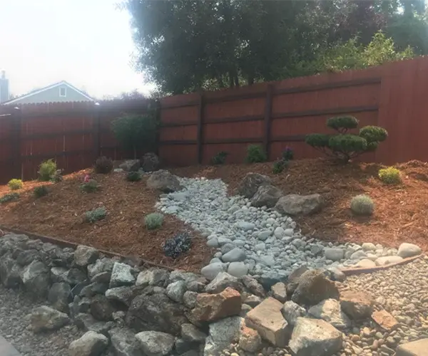 Residential landscape design featuring a sloped garden with rocks, decorative plants, and a gravel pathway, surrounded by a wooden fence, showcasing premium landscaping services in Loomis, CA.