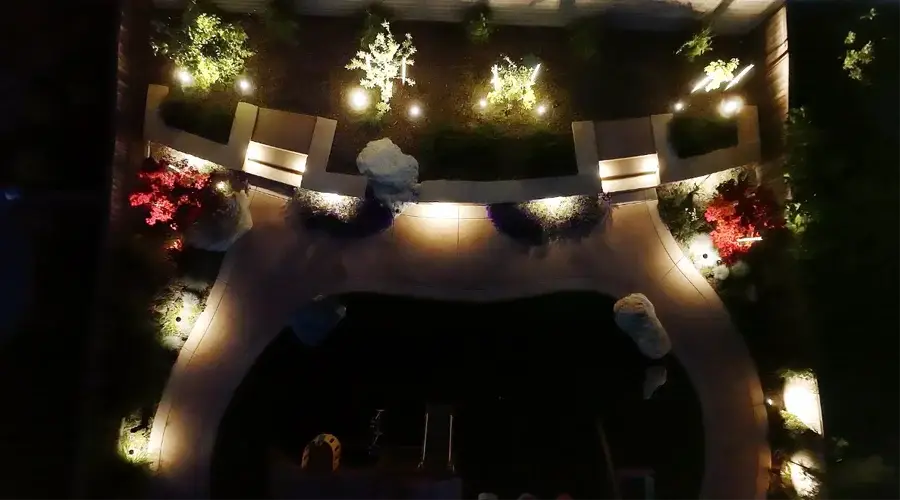 Aerial view of a beautifully landscaped outdoor space at night, featuring strategically placed path lights, uplights illuminating trees and plants, and accent lighting highlighting colorful flower beds, creating a warm and inviting ambiance for safety and aesthetics.