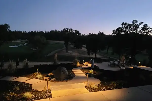 Landscape lighting illuminating a stone pathway and seating area in a serene outdoor setting at dusk, showcasing the beauty and safety enhancements provided by Rogers Landscape in Rocklin, CA.