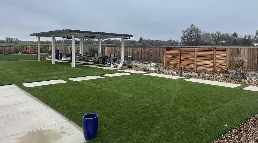 Low-maintenance artificial turf landscape featuring a covered patio area, stone pathways, and drought-tolerant plants, illustrating sustainable landscaping solutions by Rogers Landscape.