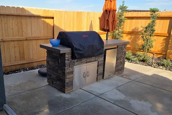 Outdoor kitchen with stone structure, built-in grill covered with black cover, stainless steel storage, and blue bowl on counter, surrounded by wooden fence and greenery, reflecting Rogers Landscape's outdoor living services in Rocklin, CA.