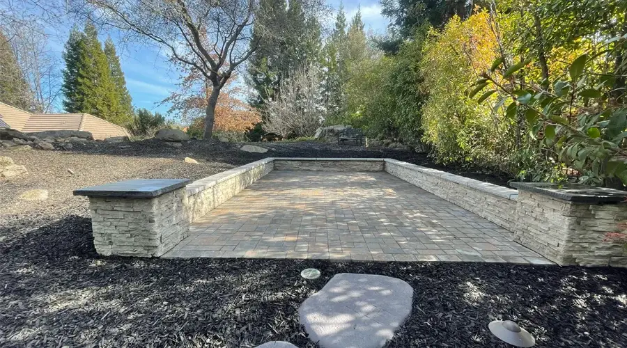 Planter wall made of stone with a paved surface in a landscaped outdoor setting, surrounded by greenery and mulch, enhancing the outdoor living space.