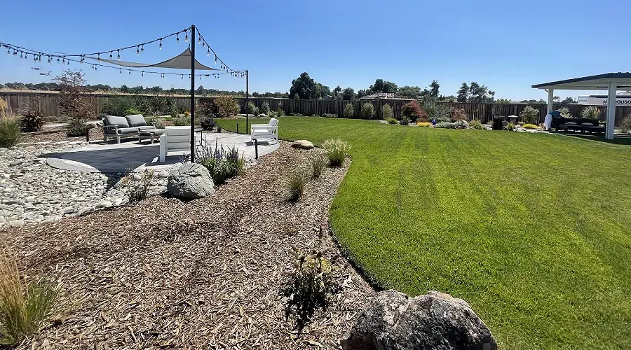 Lush green artificial turf lawn with low-water landscaping, featuring decorative rocks, native plants, and a shaded seating area, illustrating sustainable outdoor living by Rogers Landscape.