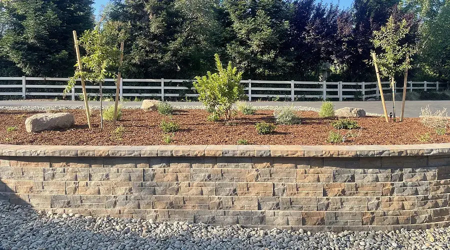 Retaining wall landscape design featuring a stone wall with various plants and trees, surrounded by decorative gravel and a white fence, illustrating professional landscaping services by Rogers Landscape.
