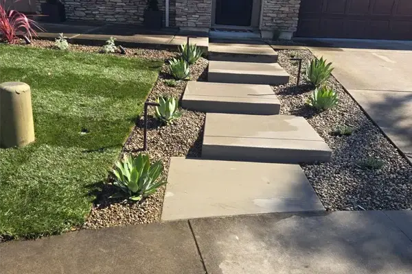 Concrete stepping stones leading to a front entrance, surrounded by decorative gravel and drought-tolerant plants, enhancing the hardscape design for a functional and visually appealing landscape.