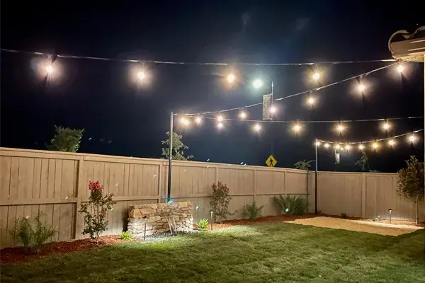 Outdoor lighting design featuring string lights illuminating a landscaped backyard at night, highlighting a stone feature and surrounding plants, enhancing ambiance and safety.