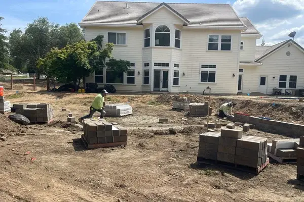 Workers laying concrete blocks for a landscape renovation project in a residential yard, with a two-story house in the background.