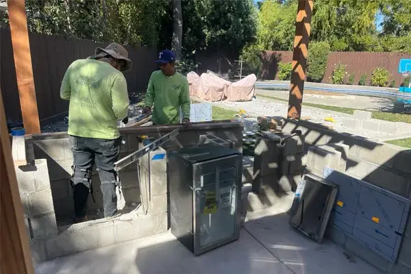 Two workers installing a custom outdoor kitchen structure, featuring a metal cabinet and preparation space, in a residential landscaping setting.