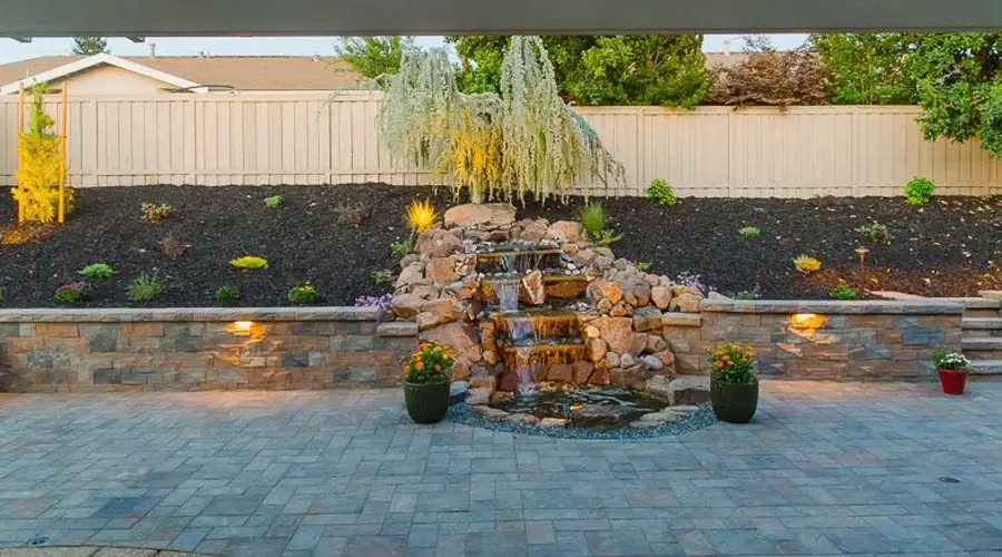 Custom stone waterfall feature in landscaped outdoor space, surrounded by plants and lighting, enhancing tranquility and visual appeal for outdoor living.