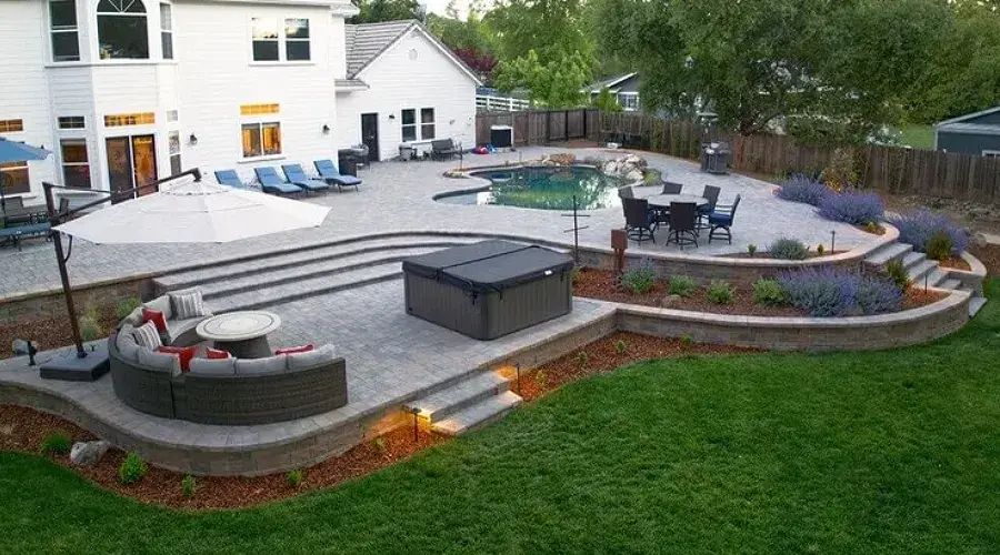 Backyard landscape featuring a stone patio with seating area, hot tub, and pool, surrounded by native plants and decorative lighting, illustrating sustainable outdoor design principles by Rogers Landscape.
