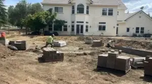 Construction site for landscape renovation featuring stacked stones and a worker, with a residential home in the background.